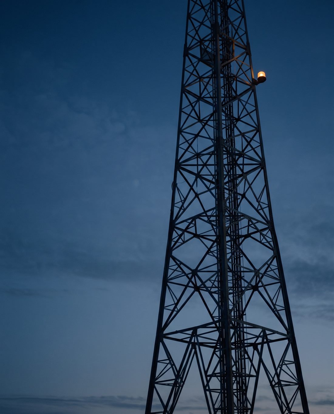 A lattice communications mast silhouetted against a deep blue dusk sky, an amber warning light glowing near the top.