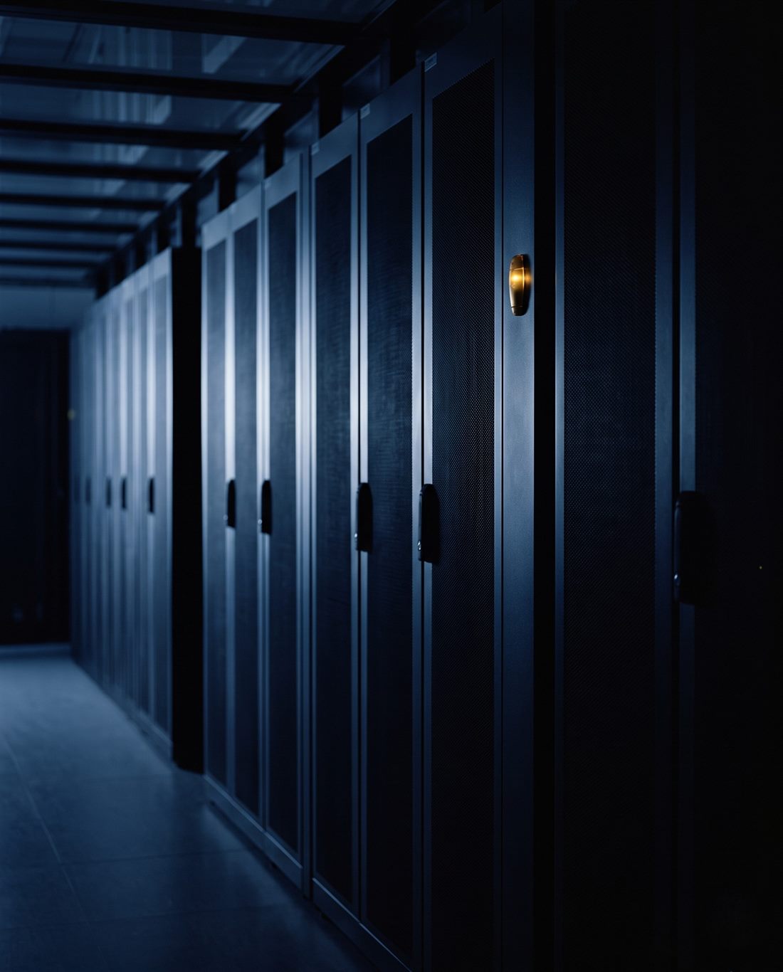 A dimly lit data centre aisle, rows of server cabinets receding into darkness with a single amber status indicator lit.